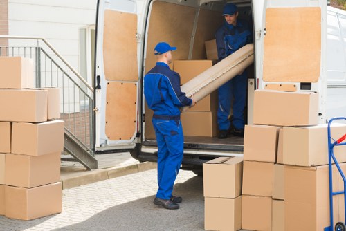 Two removal workers wearing blue uniforms and matching caps are engaged in loading a white moving van with cardboard boxes. One worker, standing inside the van, is passing down a large rolled-up item wrapped in brown paper to a second worker standing outside on the pavement, near a small staircase leading to a residential building. The van's rear doors are open, revealing an interior lined with wooden panels, ready for further packing. Several stacked cardboard boxes, in various sizes and natural brown tones, are positioned both inside and outside the van, on the ground, and on a blue trolley nearby. The setting appears to be a residential driveway or street, with a clean, paved surface. The overall environment is well-lit, possibly in daylight, and the scene captures a typical house removal operation, with furniture and belongings organized for transportation, consistent with professional removals services in Watford or nearby areas.